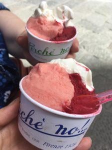 Two hands holding cups of bright pink, deep red, and white gelato, with colorful protruding spoons.