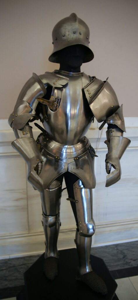 Suit of silvery plate armor, standing erect. It is undecorated and practical, with very hefty protection on the shoulders. The different and inferior time - pocked quality of the helmet displayed with it shows that it is a substitute, not part of the original surviving suit.